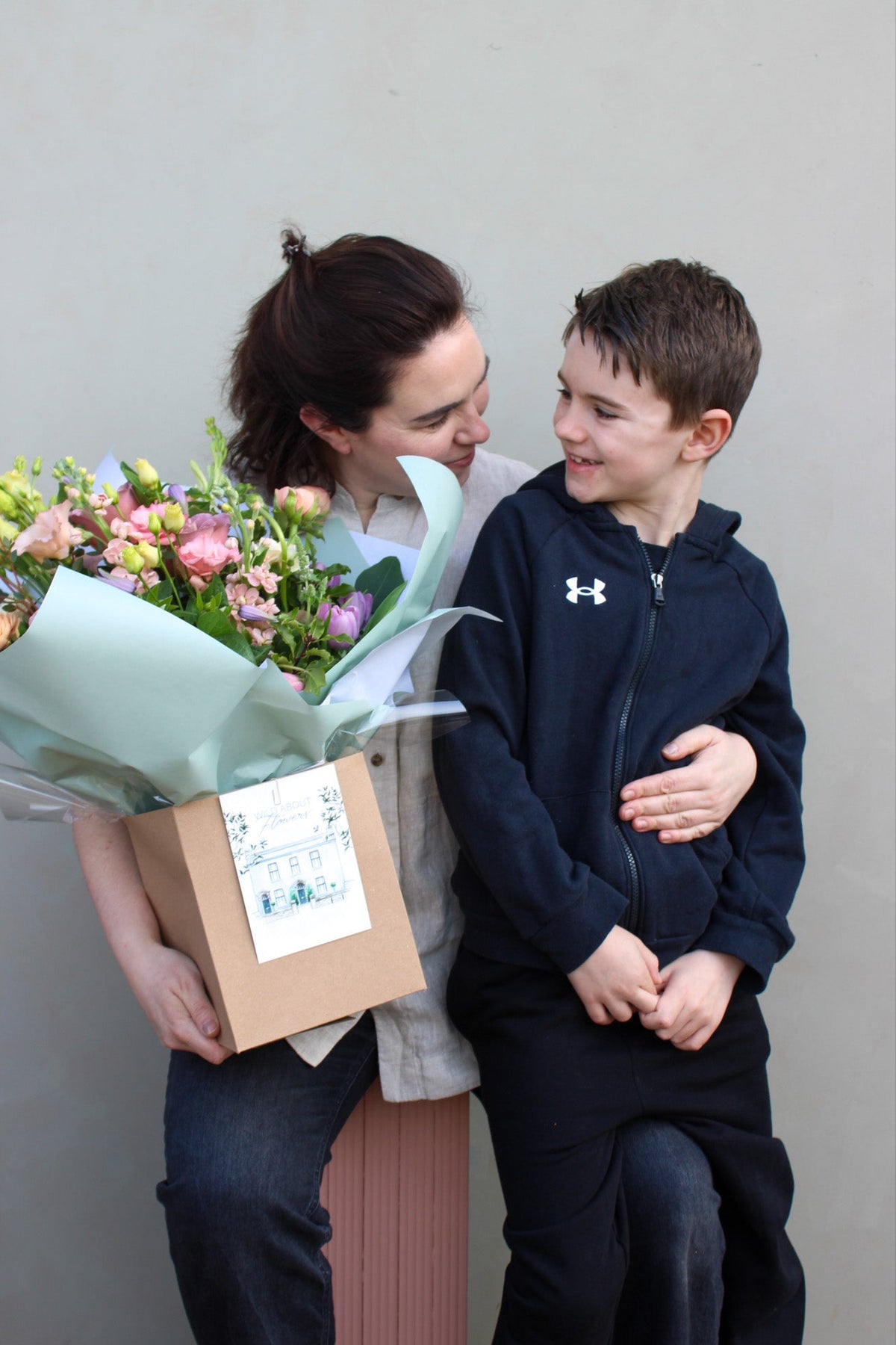 A woman on a bench holds a Secret Garden luxury bouquet from Wild About Flowers in a brown paper bag, hugging a smiling boy in a dark hoodie. They share a happy moment against a plain background.