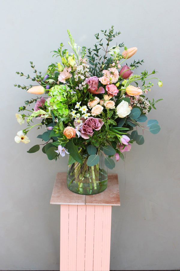 The Mother's Day Vase by Wild About Flowers features a vibrant mix of roses, tulips, and seasonal stems in a recycled glass vase, elegantly displayed on a pink wooden pedestal against a simple gray backdrop.