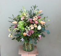 The Wild About Flowers Mother's Day Vase is a large floral arrangement in a recycled glass vase featuring seasonal flowers like pastel roses, tulips, and hydrangeas with greenery, all on a small wooden stand against a neutral background.