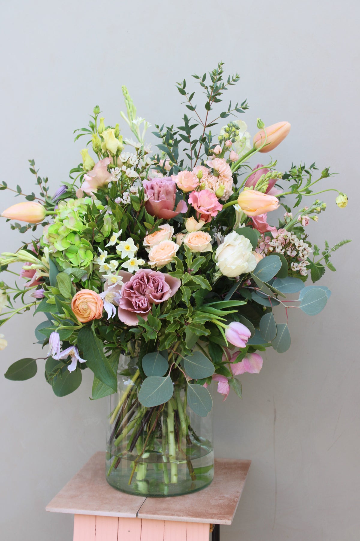 A vibrant bouquet by Wild About Flowers rests in a recycled glass Mother's Day Vase on wood. It includes pink roses, pale yellow tulips, and green hydrangeas with eucalyptus leaves, set against a neutral background—ideal for a Mother's Day centerpiece.