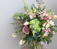 The Mother's Day Vase by Wild About Flowers is a vibrant floral arrangement in a recycled glass vase featuring pink roses, green hydrangeas, yellow tulips, white chamomile, and seasonal stems set against a light backdrop.