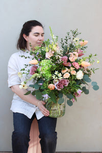 A person showcases a Wild About Flowers Mother's Day Vase, filled with a large bouquet of assorted roses, tulips, and greenery. Against a light-colored wall, their long hair and attire of a light shirt and dark pants enhance the scene's serene beauty and sustainable elegance.