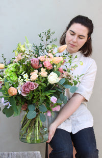 An individual in a light shirt and dark pants holds the "Mother's Day Vase" by Wild About Flowers, a large recycled glass vase filled with roses, tulips, and greenery, against a plain background.