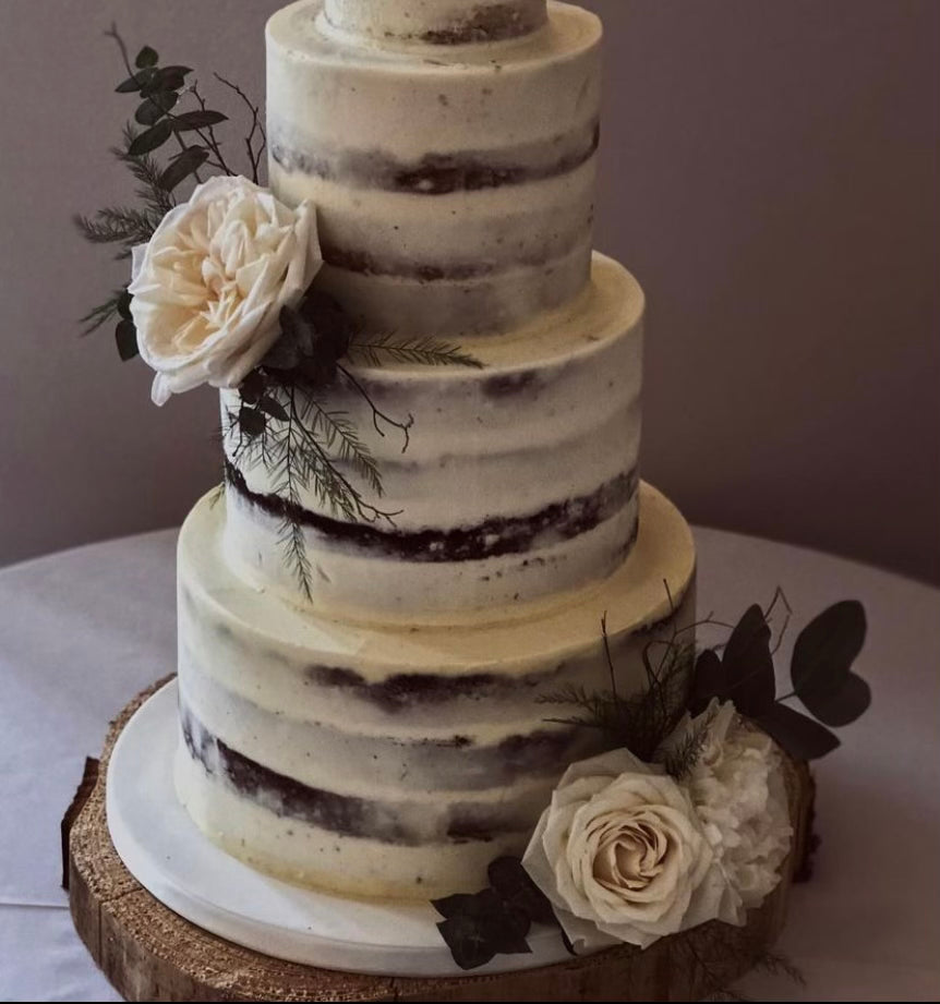 The Cake flower duo by Wild About Flowers features two large white roses, delicate foliage sprigs, and greenery. This stunning arrangement offers an exquisite accent to a three-tiered semi-naked wedding cake with elegant white frosting, beautifully resting on a rustic wooden slab.