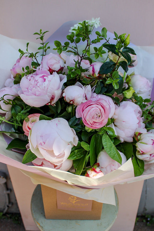 Peony Love Hand Tied by Wild About Flowers features elegant peonies and roses with greens, wrapped in white paper in a beige box atop a green stool—an exquisite floral gift set against a soft, light background.