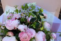 The Peony Love Hand Tied by Wild About Flowers features premium peonies and ranunculus with greenery, elegantly wrapped in white and lavender paper—an elegant floral gift set against a pale background.