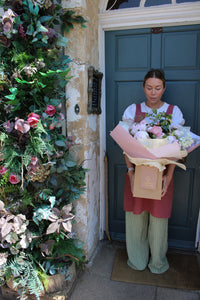 Wearing an apron, a woman stands before a blue door holding a Wild About Flowers Peony Love Hand Tied bouquet filled with premium peonies and greenery. Similar lush, colorful floral creations adorn the wall beside her.