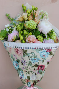 The Peony Floral Wrap by Wild About Flowers features pastel seasonal flowers, including peonies and green hydrangeas, wrapped in a limited edition summer fabric with pink and blue blooms on beige.