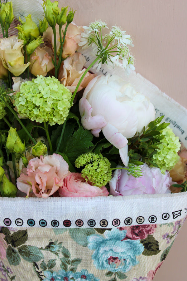 The Peony Floral Wrap by Wild About Flowers features pastel seasonal blooms—white peonies, green hydrangeas, and pink roses—wrapped in a limited edition summer fabric, displayed on a light pink background.