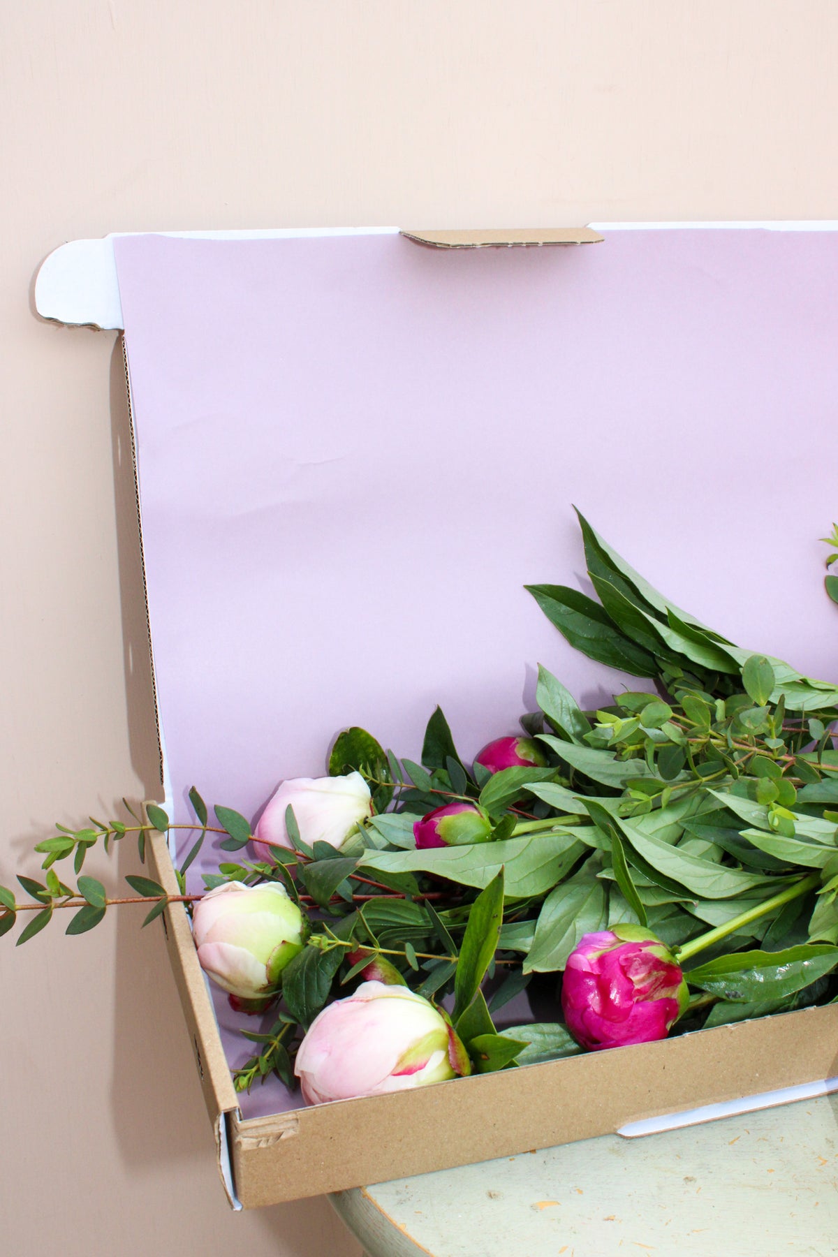 Peonies by Post from Wild About Flowers features pink and white peonies with green foliage in a light purple-lined cardboard box, designed letterbox-friendly for easy delivery.