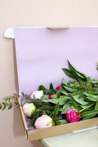 Peonies by Post from Wild About Flowers features pink and white peonies with green foliage in a light purple-lined cardboard box, designed letterbox-friendly for easy delivery.