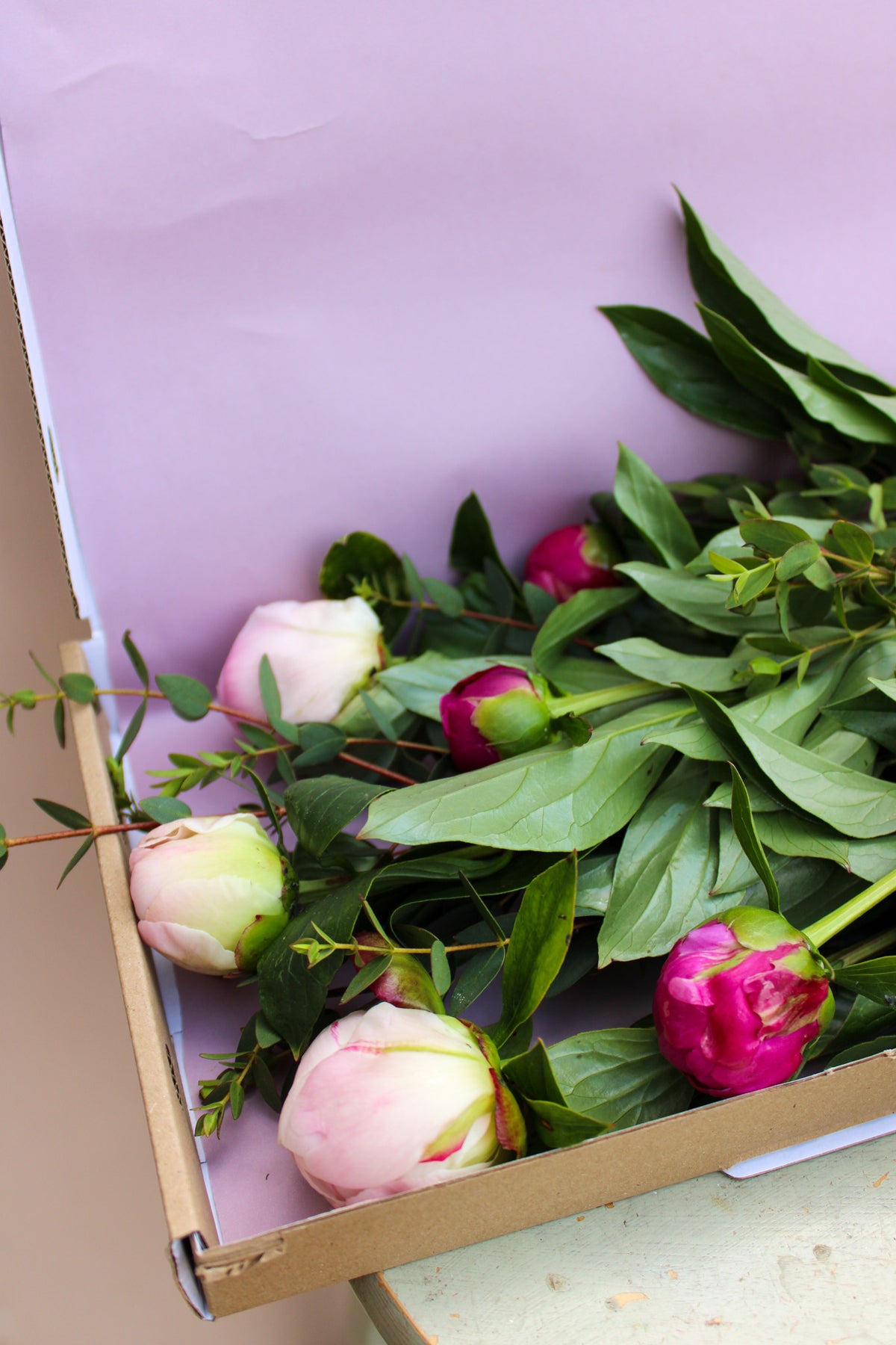 Peonies by Post from Wild About Flowers features fresh pink and magenta peony stems with lush green leaves, delivered in a letterbox-friendly cardboard box.