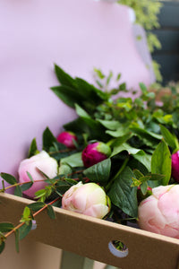 A cardboard box of fresh green leaves and pink Peonies by Post from Wild About Flowers—perfect letterbox-friendly blooms—rests against a soft purple background.