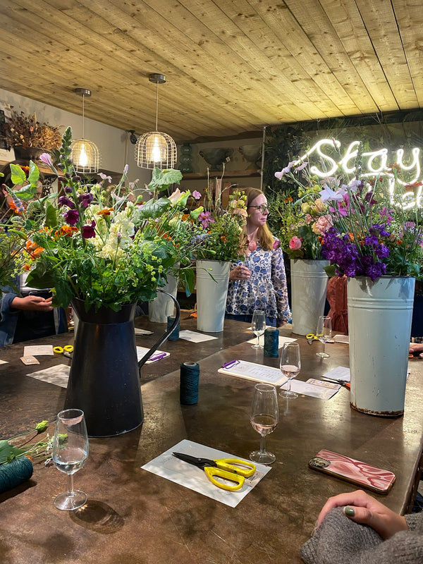 In a cozy room with a wooden ceiling, a woman teaches a floral arrangement class called Creative Souls by Event. The table is adorned with large vases of colorful flowers, scissors, twine, papers, and wine glasses. A neon "Stay" sign fosters connection in this vibrant floral shop setting.