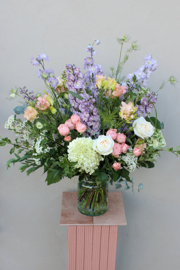 The "Gorgeous bouquet" from Wild About Flowers is an exquisite arrangement crafted by skilled florists. It features fresh flowers, including roses and delphiniums in shades of white, pink, and lavender. The bouquet is beautifully displayed in a glass vase on a small, pink pedestal against a plain, light gray background. Lush greenery is interspersed throughout for added elegance.