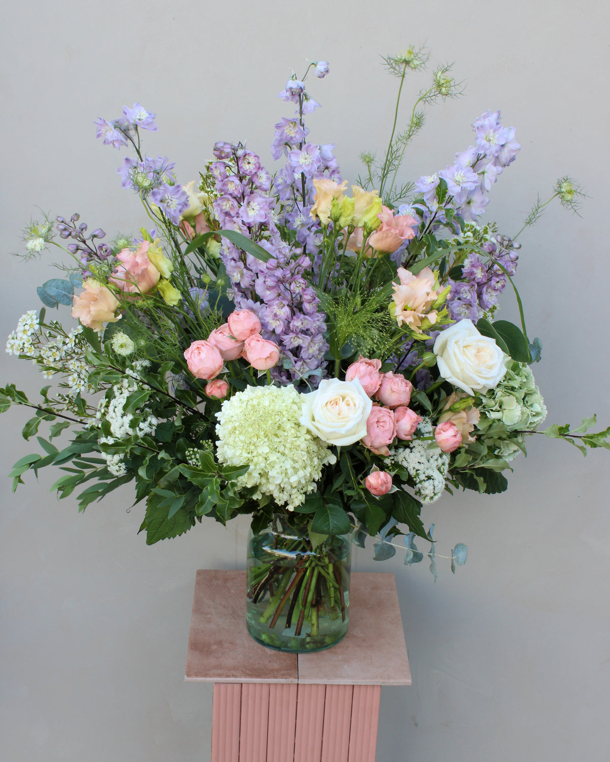 The "Gorgeous bouquet" by Wild About Flowers showcases an exquisite arrangement of white and pale pink roses, hydrangeas, lilac delphiniums, and various green foliage. Expertly curated by skilled florists, this stunning display is presented in a clear glass vase on a pink pedestal against a neutral background.