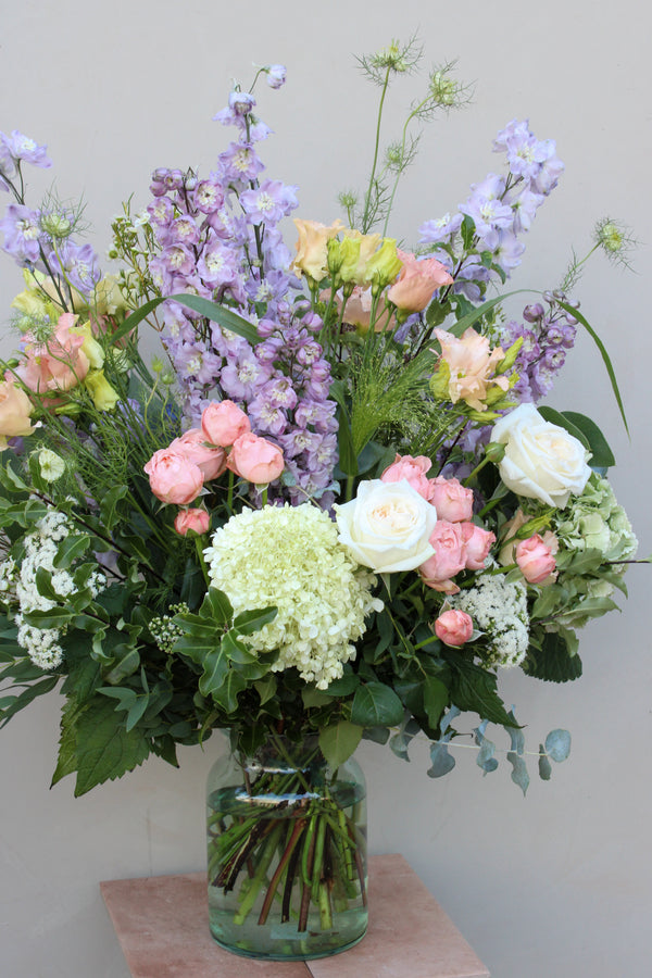 The "Gorgeous Bouquet" by Wild About Flowers features a stunning glass vase filled with white and pink roses, light purple delphiniums, white hydrangeas, and assorted greenery. Arranged by skilled florists, the fresh flowers create an abundant and lush display against a plain background.