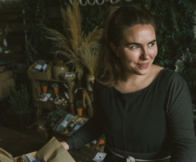Woman standing behind a shop counter, smiling, with plants and gift items displayed on shelves behind her