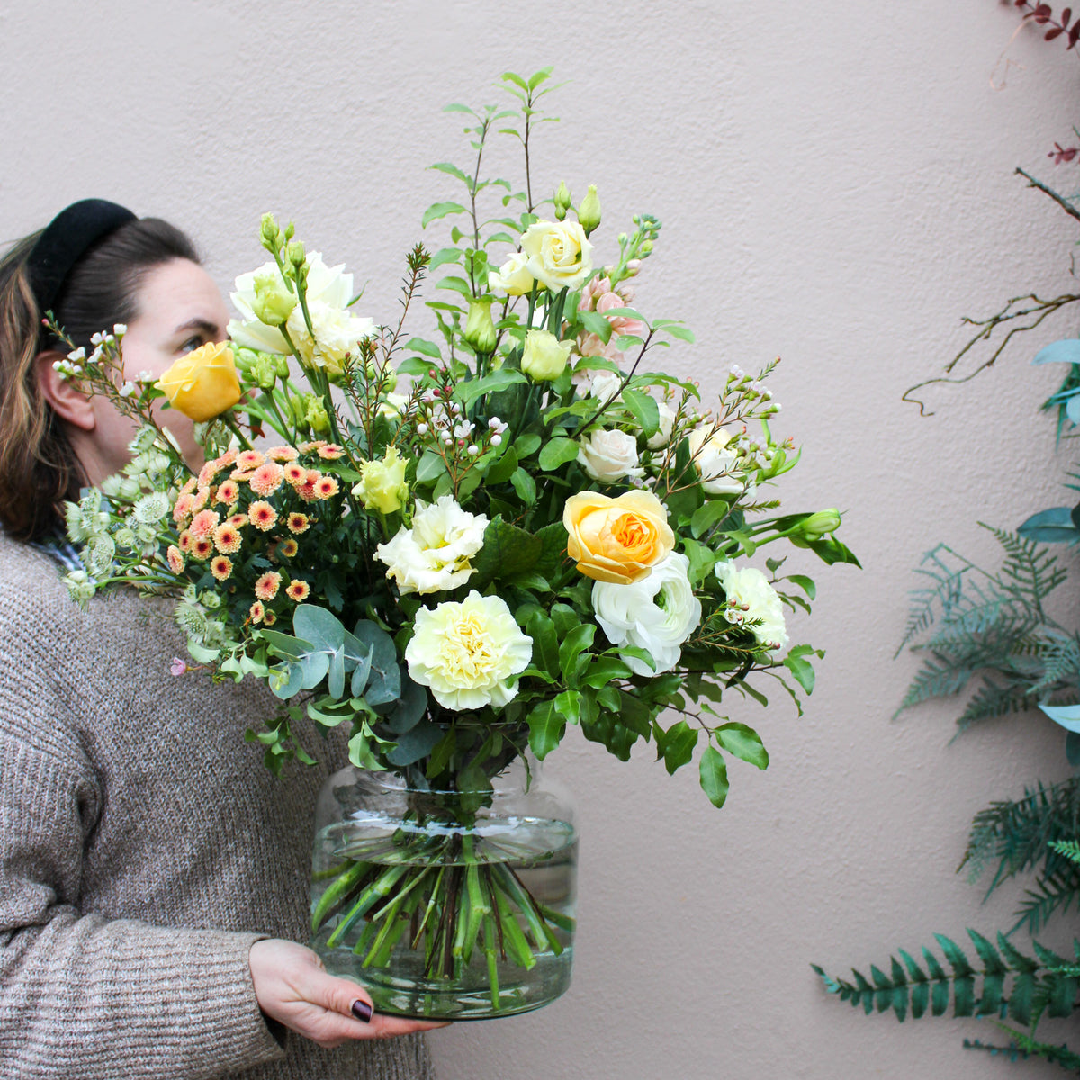 A person in a beige sweater holds the 'Jar of Joy' vase arrangement from Wild About Flowers—an elegant display of pastel roses and greenery—creating a luxurious flower gift against a light pink wall.