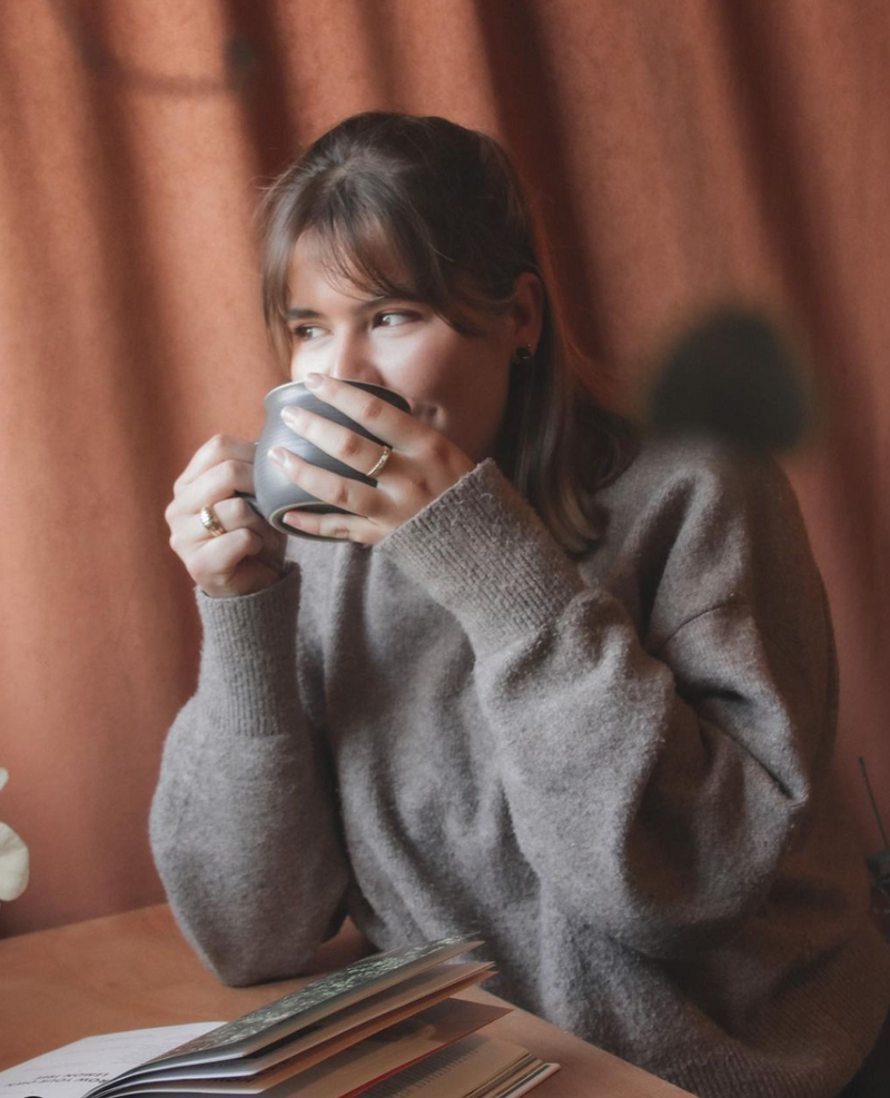 Woman in a grey sweater sitting at a table, holding a mug and looking to the side, with books in front of her