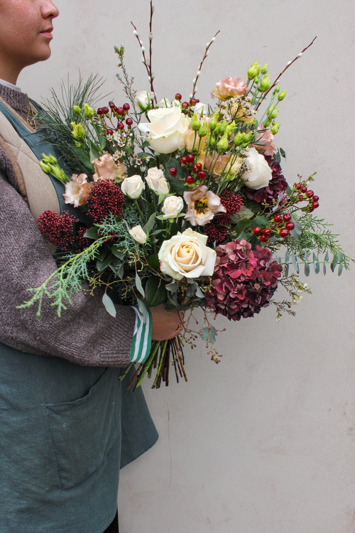 A person in a gray sweater and green apron holds the Wild About Flowers Love Actually Hand Tied—a festive bouquet of white roses, red berries, and lush greenery—perfect as Christmas flowers—against a plain light background.