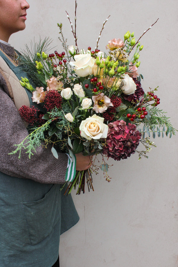 A person in a gray sweater and green apron holds the Wild About Flowers Love Actually Hand Tied—a festive bouquet of white roses, red berries, and lush greenery—perfect as Christmas flowers—against a plain light background.