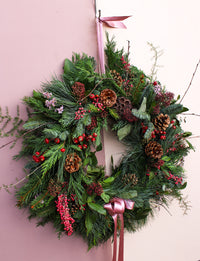 The Love Actually Winter Wreath by Wild About Flowers features pine branches, red berries, pinecones, and mixed greenery with a pink satin ribbon, beautifully displayed on a pale pink wall.