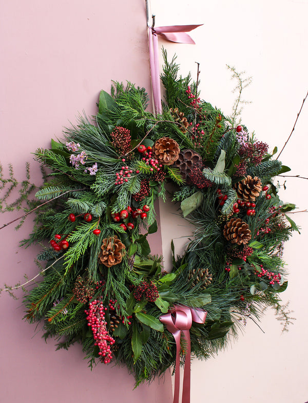 The Love Actually Winter Wreath by Wild About Flowers features pine branches, red berries, pinecones, and mixed greenery with a pink satin ribbon, beautifully displayed on a pale pink wall.