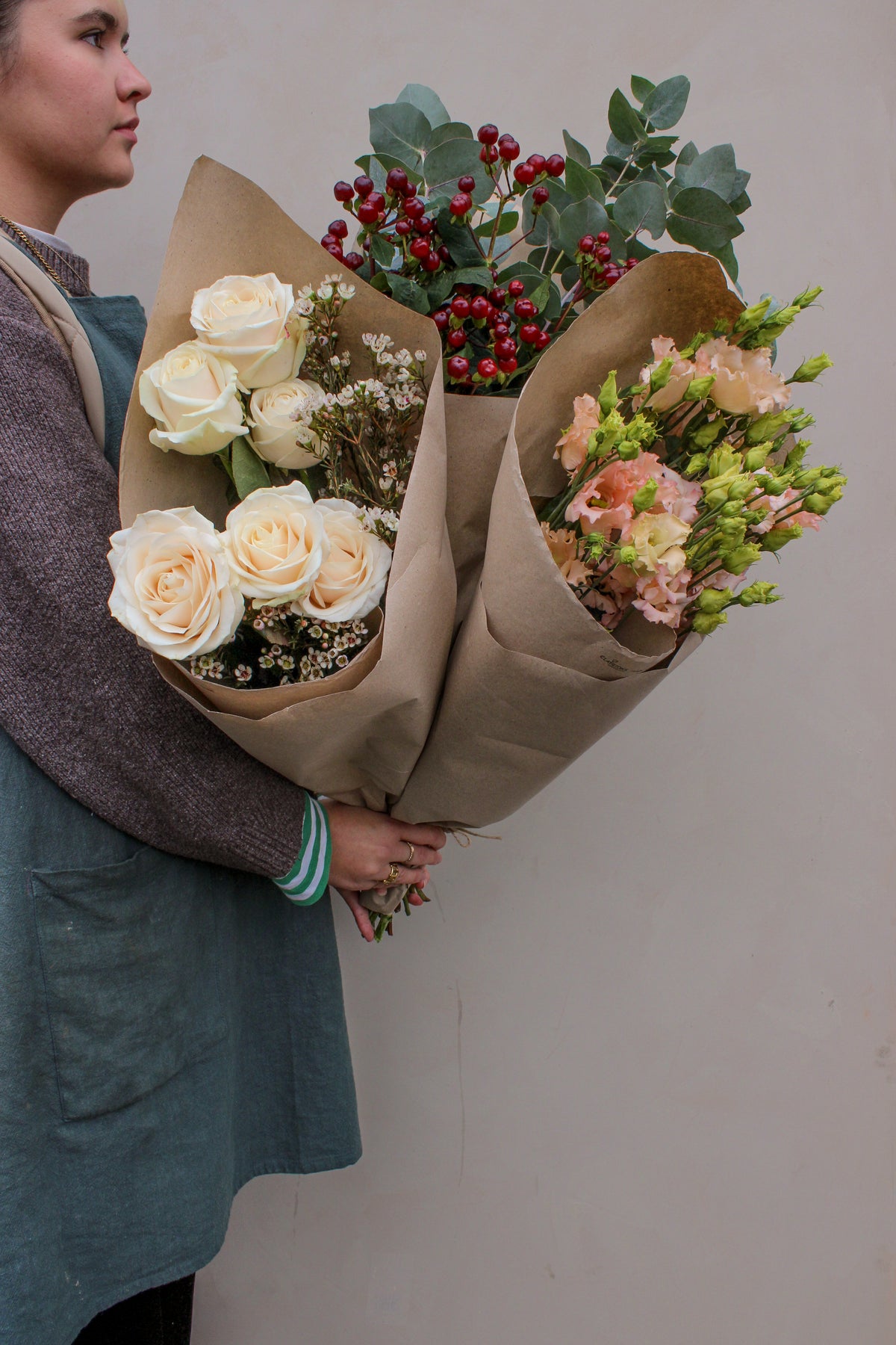 A person in a blue apron holds three Love Actually Seasonal Stems bouquets by Wild About Flowers—white roses, red berries, greenery, and peach blooms wrapped in brown paper—perfect for Christmas gifting against a beige background.