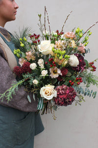 A person in a brown sweater and green apron holds a stunning Love Actually Hand Tied by Wild About Flowers, featuring mixed blooms—perfect for flower delivery Keynsham—set against a plain light background.