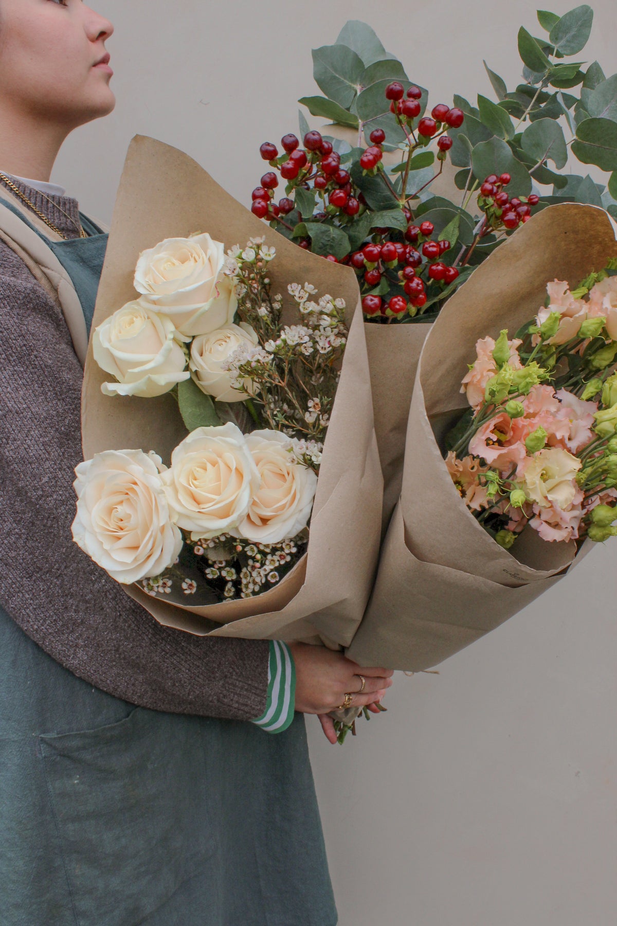 A person holds three large Love Actually Seasonal Stems bouquets by Wild About Flowers, with white roses, red berries, greenery, and peach seasonal blooms wrapped in brown paper, set against a plain light background.