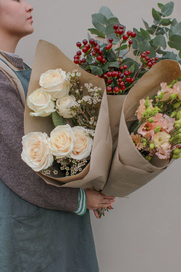 A person holds three large Love Actually Seasonal Stems bouquets by Wild About Flowers, with white roses, red berries, greenery, and peach seasonal blooms wrapped in brown paper, set against a plain light background.