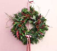 The Wild About Flowers Love Actually Winter Wreath, crafted with pine, red berries, leaves, pinecones, and a pink ribbon bow, brings charming seasonal style to any space.