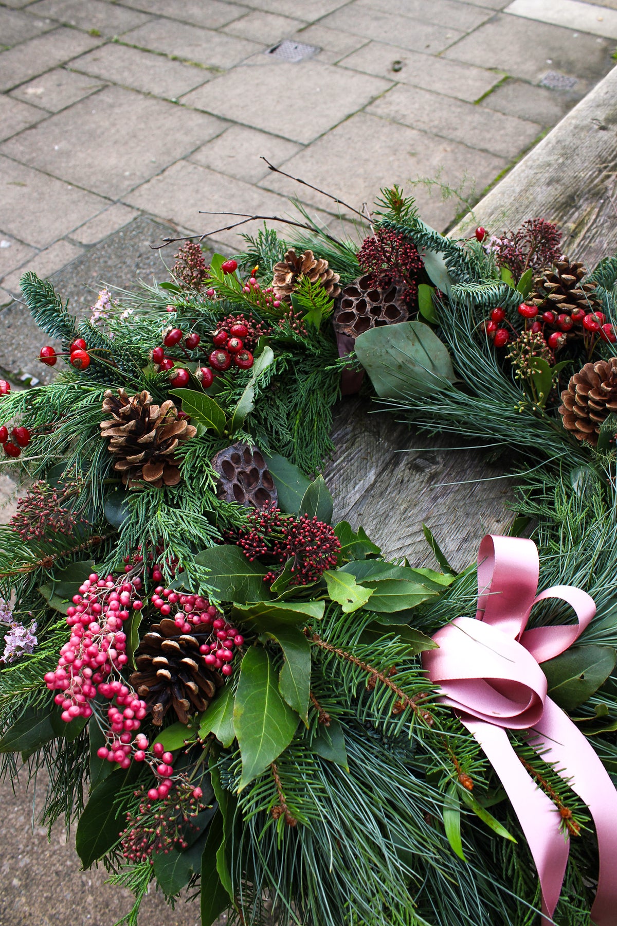 The Love Actually Winter Wreath by Wild About Flowers features handcrafted pine branches, pine cones, red berries, greenery, and a pink ribbon—perfect for winter décor.
