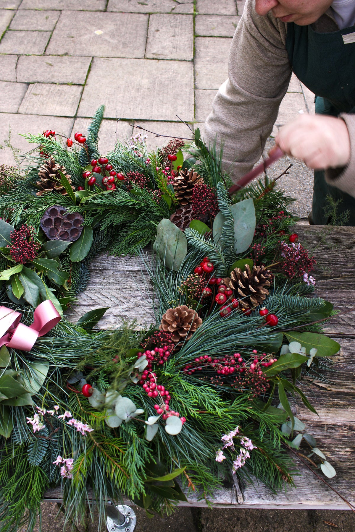 A person arranges the Love Actually Winter Wreath by Wild About Flowers, handcrafted with pine branches, pinecones, red berries, and green foliage, on a rustic wooden outdoor table.