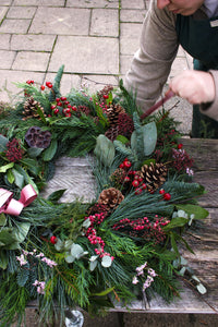 A person arranges the Love Actually Winter Wreath by Wild About Flowers, handcrafted with pine branches, pinecones, red berries, and green foliage, on a rustic wooden outdoor table.