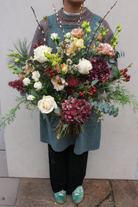 A person in a gray sweater, blue apron, and green sneakers holds the Wild About Flowers Love Actually Hand Tied bouquet—white roses, hydrangeas, berries, and greenery—ideal for Keynsham flower delivery, standing on stone with a light background.