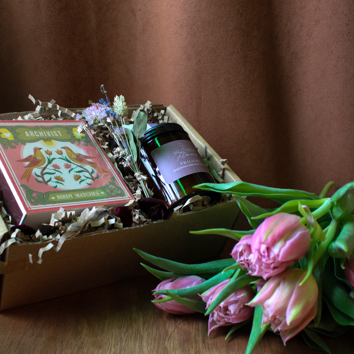 Archivist matches, candles, dried florals in a gift box with and pink tulips on a wooden surface with a brown background