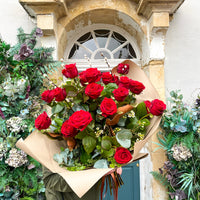 A person holds Wild About Flowers' 'The Ultimate Valentine' Red Rose Bouquet, featuring luxury red roses and greenery wrapped in brown paper, standing before a doorway decorated with lush plants and blooms.