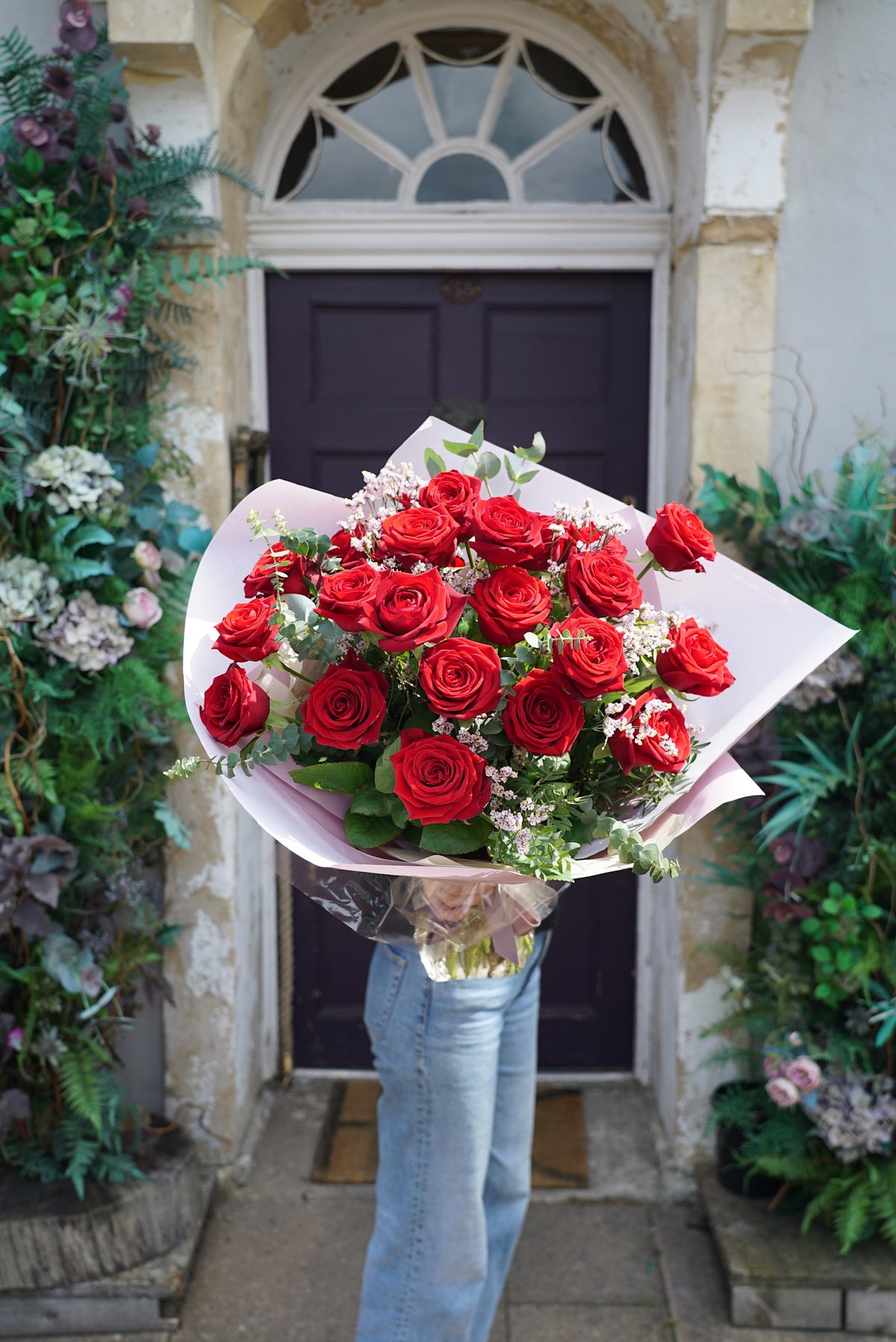 A person in blue jeans holds Wild About Flowers' 'The Ultimate Valentine' Red Rose Bouquet—vivid red roses with white fillers, wrapped in white paper—standing before a dark purple door surrounded by lush greenery.