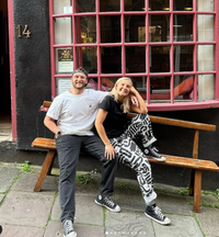 Two people sit on a bench in front of a building with large, pink-framed windows, wearing casual clothes and sneakers. One sports geometric patterned pants. Both smile at the camera, perhaps enjoying the atmosphere of the Supper Club with Cin Cin event.