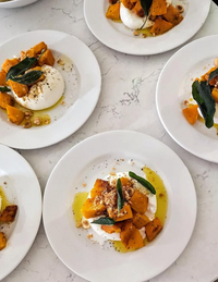 On a white countertop, "Supper Club with Cin Cin" by Event showcases plates of burrata with roasted butternut squash, sage leaves, olive oil drizzle, garnished with ground pepper and nuts.