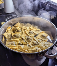 A pot of Supper Club with Cin Cin ravioli from Event is boiling on the stovetop, with pasta bubbling in the water and steam rising. The square-shaped ravioli appears partially cooked, some floating on the surface.