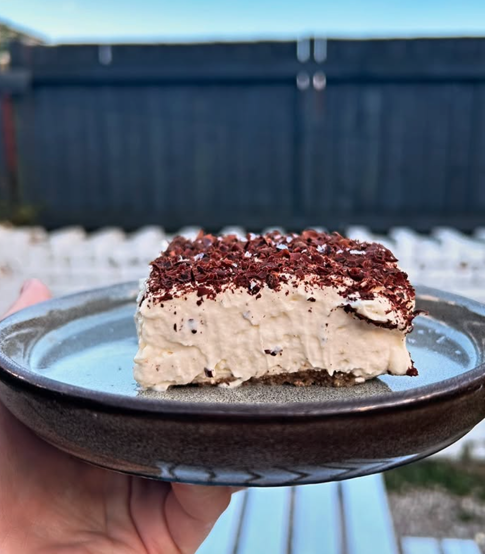 A hand holds a creamy dessert topped with chocolate shavings on a brown ceramic plate from the Supper Club with Cin Cin by Event. The background reveals a blurred outdoor setting featuring a dark fence and white seating area.