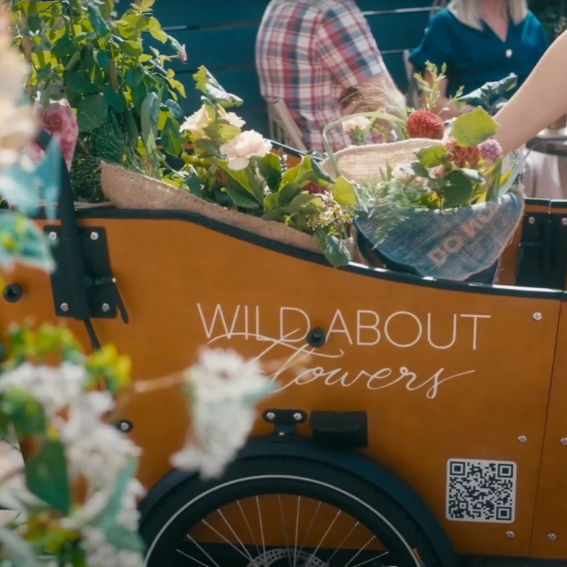 Orange bike trailer with 'Wild About Flowers' branding and a QR code, filled with flowers and plants.