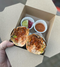 A hand holds an open Summer Sessions takeout box by Event with a halved burrito showing rice, beans, and sauce, plus sides of salsa, pickled onions, and a lime wedge from the Mexican food pop-up.