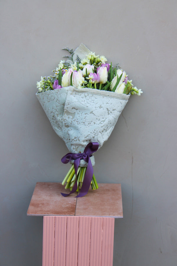 The Spring Cheer Bunch by Wild About Flowers showcases fresh white and pink tulips elegantly wrapped in patterned paper with a purple ribbon, beautifully displayed on a pink pedestal against a plain gray backdrop, adding sustainable style to any setting.