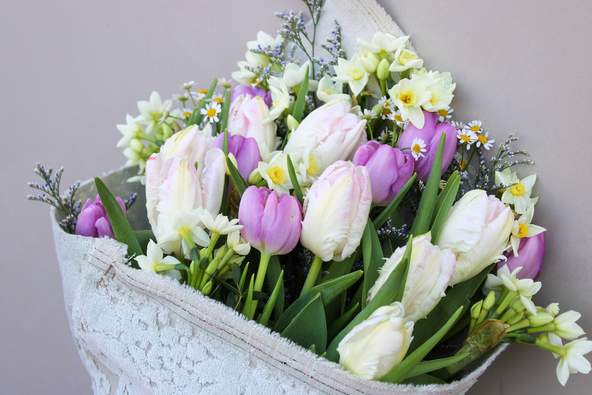 Presenting the Spring Cheer Bunch by Wild About Flowers: elegant light pink and purple tulips with white daffodils and yellow-centered daisies, wrapped in textured white paper. A radiant, sustainable bouquet to brighten your day.