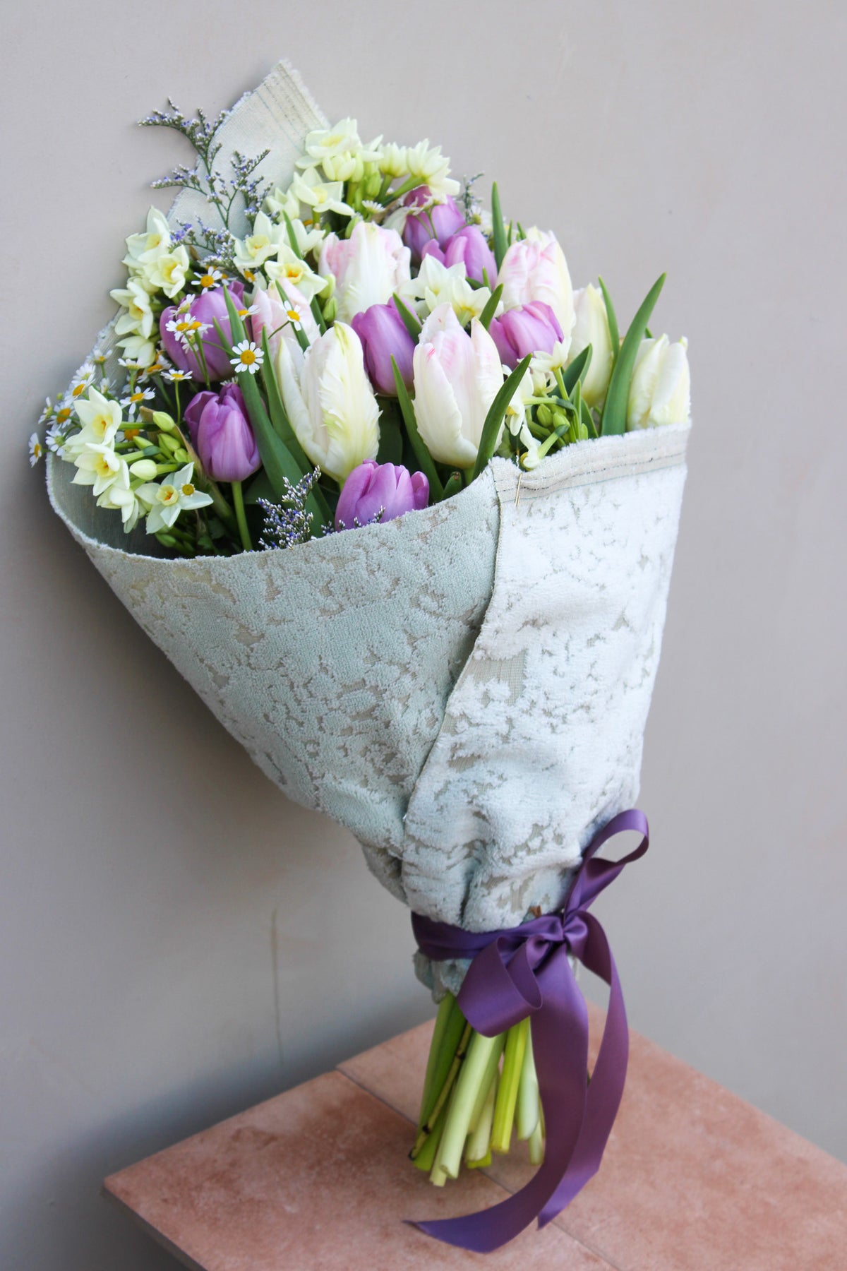 The Spring Cheer Bunch by Wild About Flowers includes seasonal tulips, white daffodils, and greenery wrapped in up-cycled fabric with a purple ribbon, beautifully leaning against a gray wall on a small pink table.