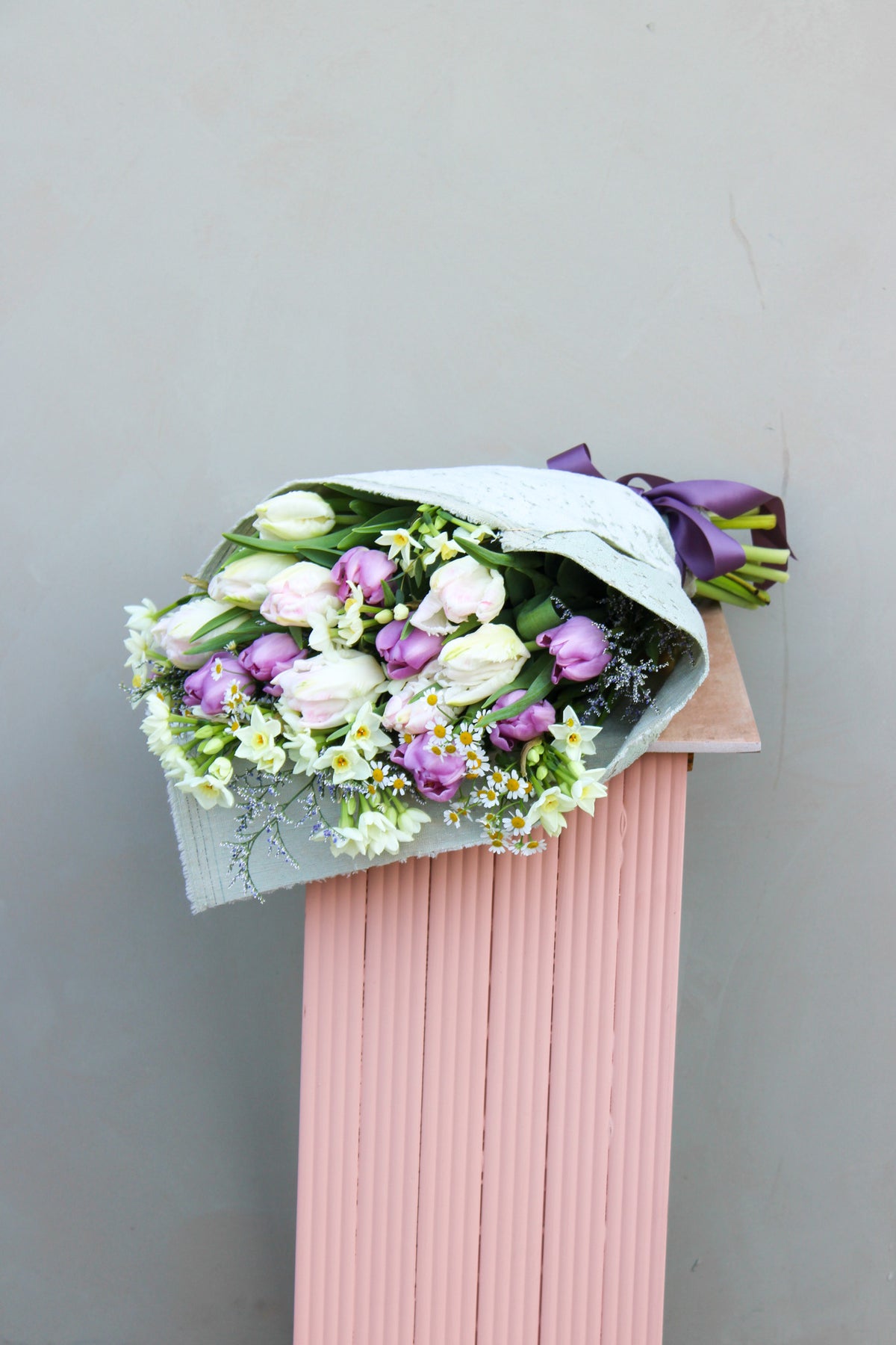 Wild About Flowers' Spring Cheer Bunch displays white and purple seasonal tulips, wrapped in up-cycled fabric with a purple ribbon, elegantly positioned on a pink column against a gray background.